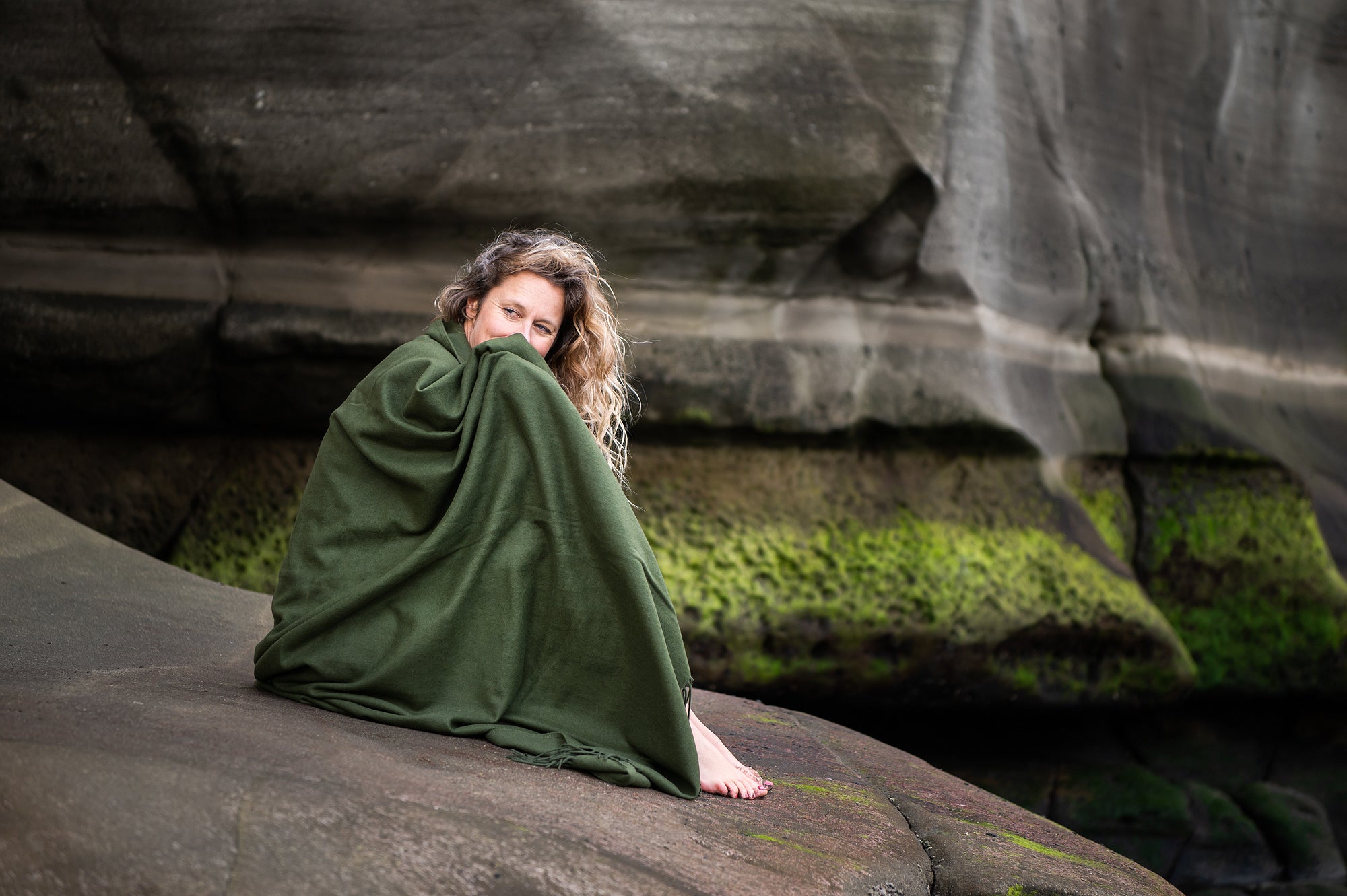 Woman sitting on rocks wrapped in cashmere merino blanket in Moss green