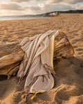 A woven beach towel with fringed edges draped over a piece of driftwood on a sandy beach, with the ocean and cloudy sky in the background.